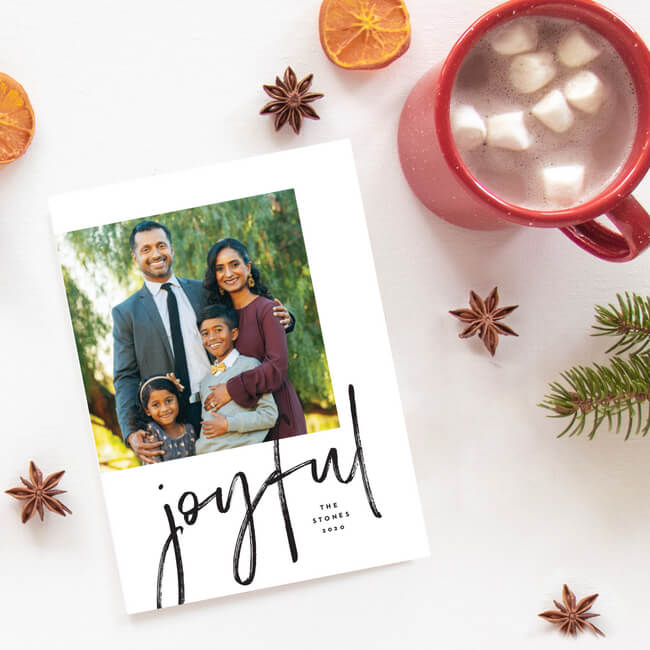 simple elegant christmas photo card featuring a smiling family. sits on a white table next to a cup of hot cocoa