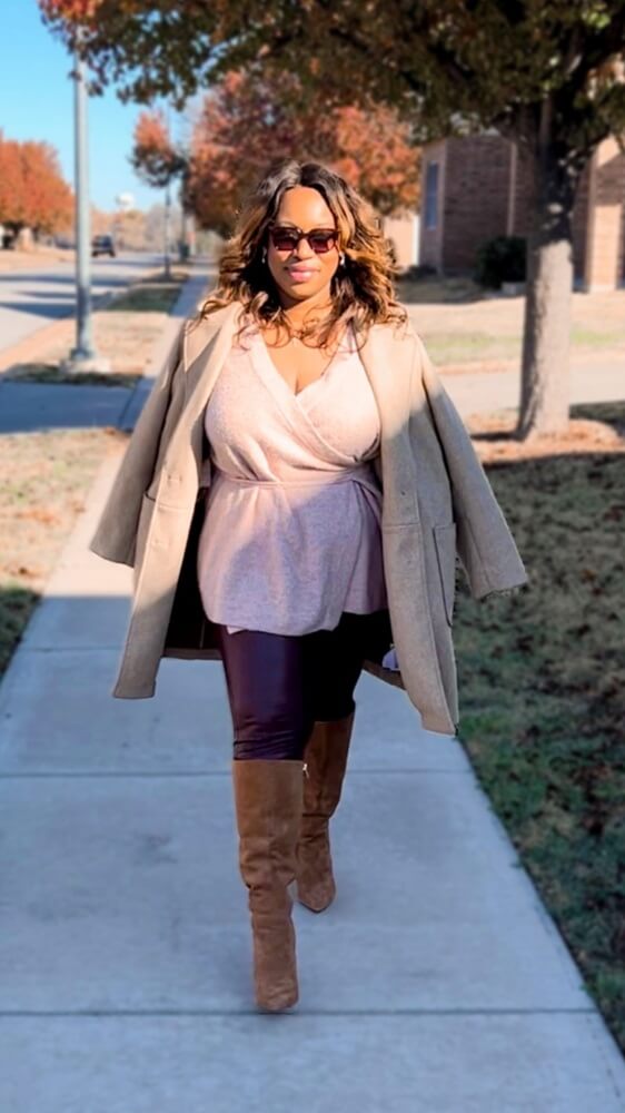 black woman walking towards the camera wearing suede knee high boots, leather leather leggings, pink sweater, tan overcoat