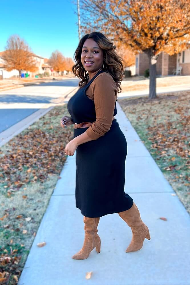 smiling woman wearing brown long-sleeved shirt under belted black tank midi length dress with brown suede knee high boots
