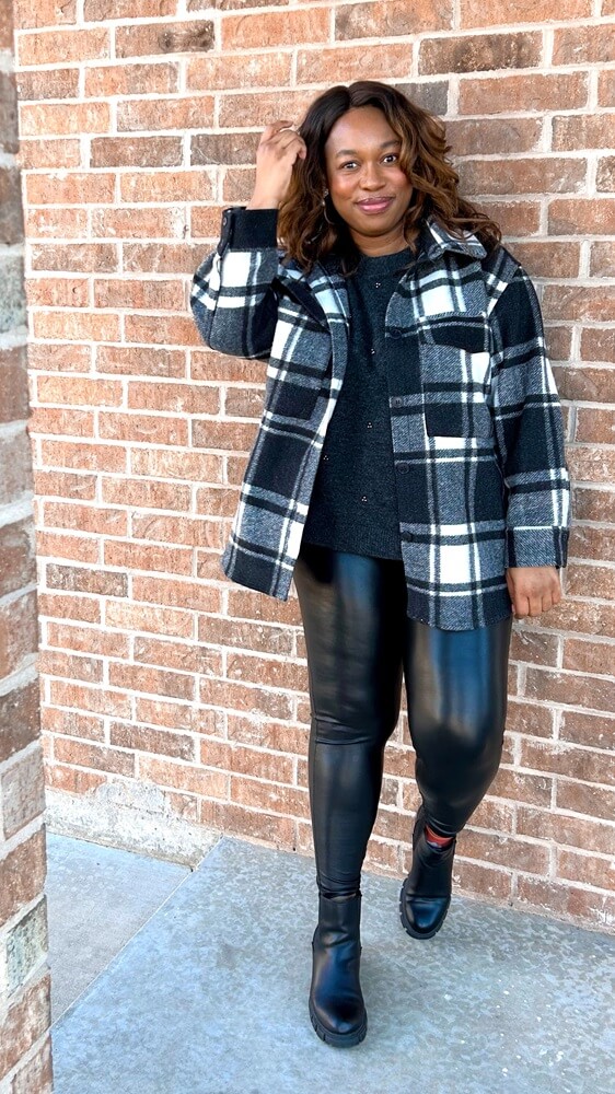 brown skin lady smiles at the camera wearing cute leather leggings outfit with dark grey sweater and black and white plaid shacket 