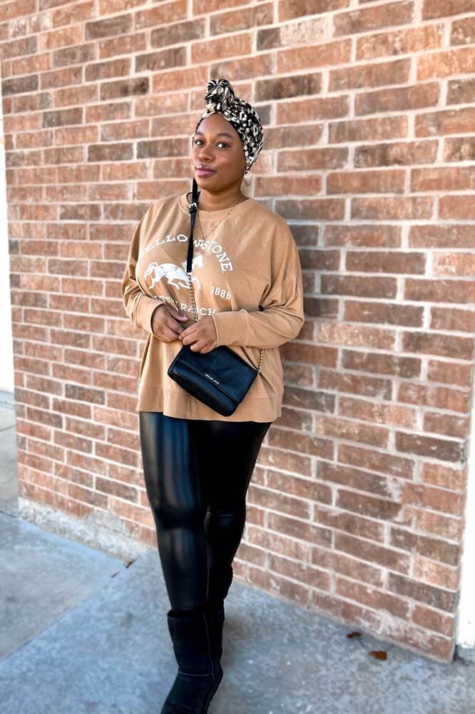 black woman carrying a black crossbody bag, wearing a brown "yellowstone" sweatshirt and black and taupe turban style pre knotted headwrap