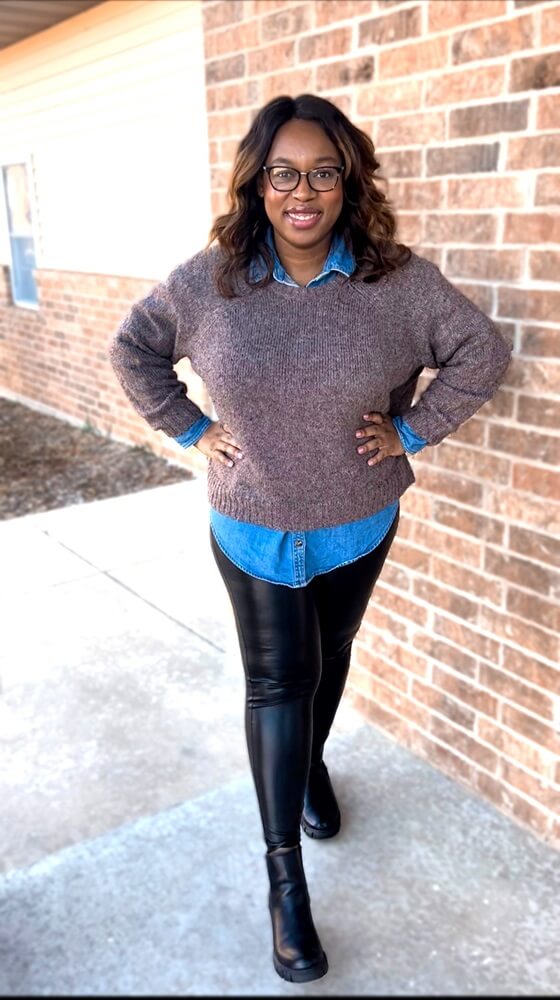 smiling woman wearing faux leather leggings and a brown sweater layered over a denim top