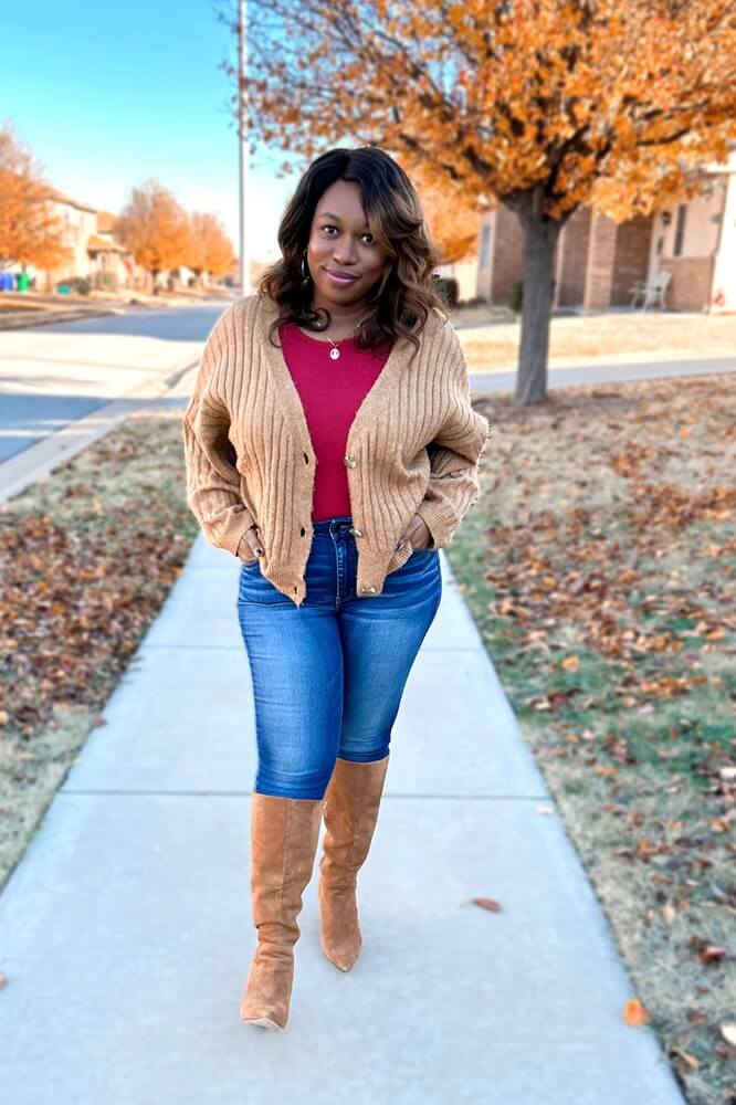 smiling Black woman wearing a brown cropped cardigan, solid burgundy top, and jeans with knee high boots